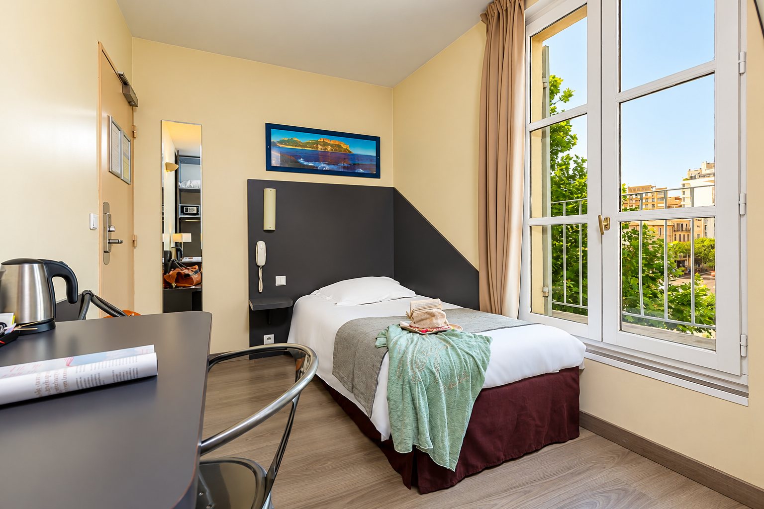 Chambre d'hôtel lumineuse avec un lit simple, un bureau et une grande fenêtre sur les arbres